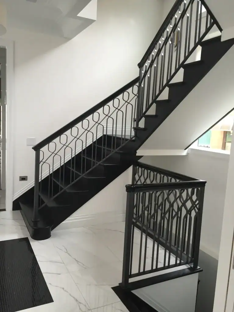 A modern indoor staircase with black railings and geometric patterns, set against white walls and glossy white marble floors. Natural light brightens the clean, minimalist space.